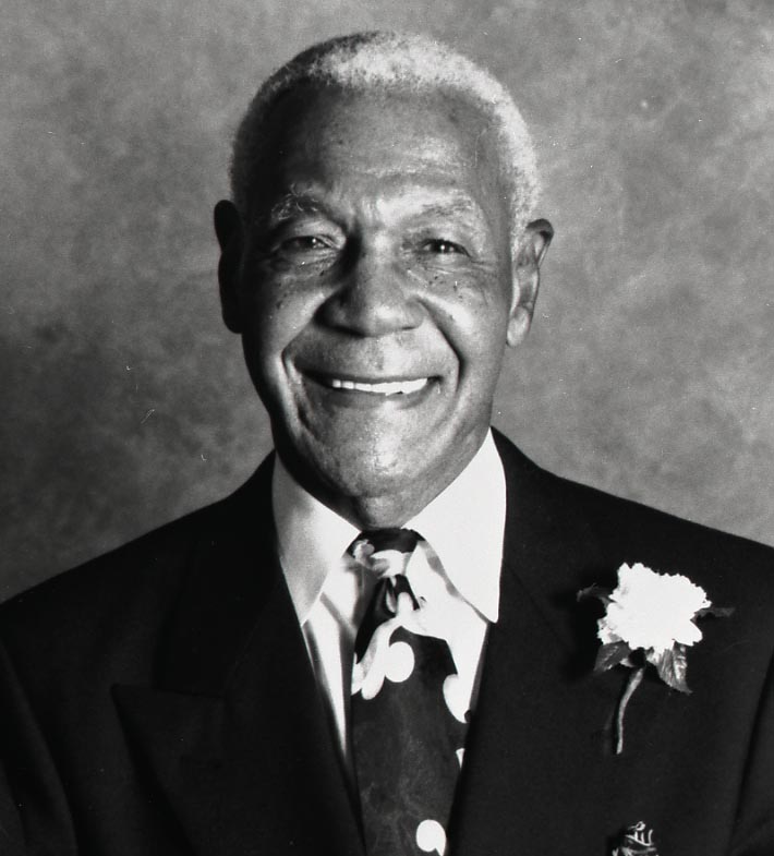 Smiling elderly man in a suit with a floral tie and boutonniere on a plain backdrop.