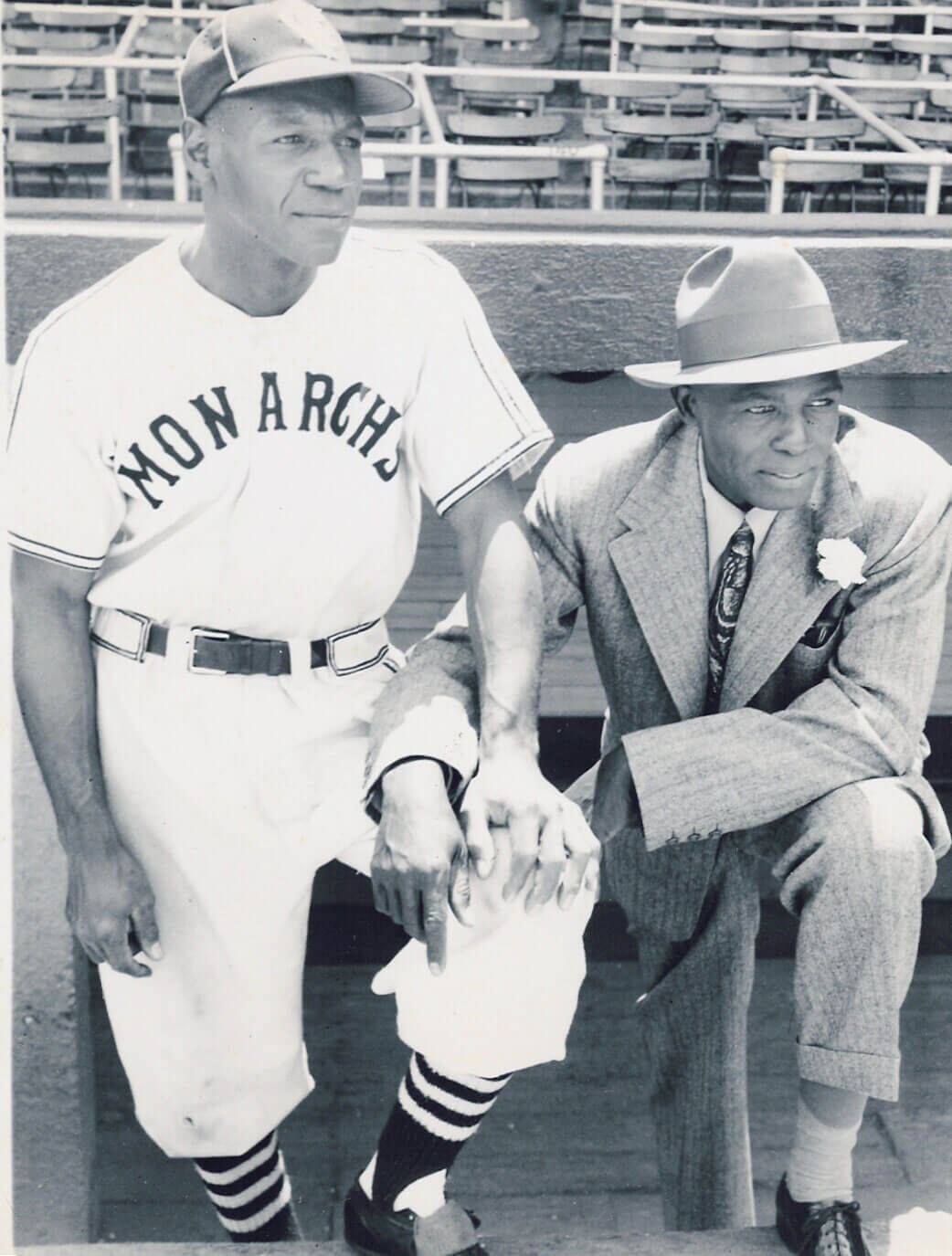Two men pose in an empty baseball stadium. One wears a MONARCHS baseball uniform and cap, standing and resting his hand on a bat. The other is beside him kneeling, dressed in a suit and hat, looking off to the side.