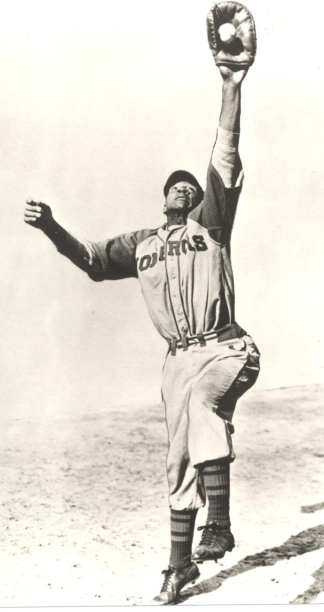 A baseball player in a vintage uniform jumps high with an outstretched glove to catch a ball. He wears a cap and striped socks, and the ground appears sandy. The photograph is in black and white.