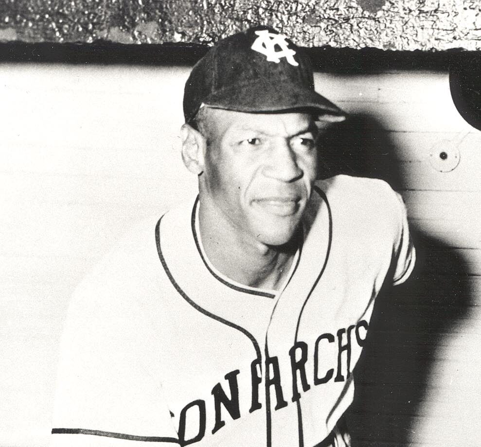 A black and white photo of a baseball player wearing a Kansas City Monarchs uniform and cap. He appears to be leaning slightly, looking off-camera. The uniform and cap have the team’s initials and name visible.