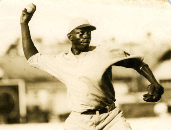 A vintage photo of a baseball player in mid-pitching action. Hes wearing a classic uniform and cap, with a focused expression. The background shows a blurred field and stands, suggesting an in-game scene.