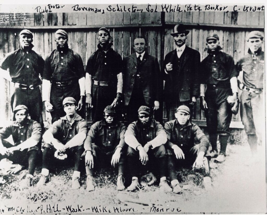 A vintage black and white photo of a baseball team. Eleven men pose, some standing and some seated, wearing dark uniforms and caps. A wooden fence serves as the backdrop. Handwritten names are visible on the image.