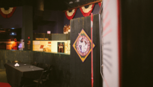 A dimly lit room features a black table with chairs and a backdrop of dark walls adorned with vintage-style baseball memorabilia. A circular sign with a baseball player graphic is prominently displayed. Red and white bunting decorates the upper section.