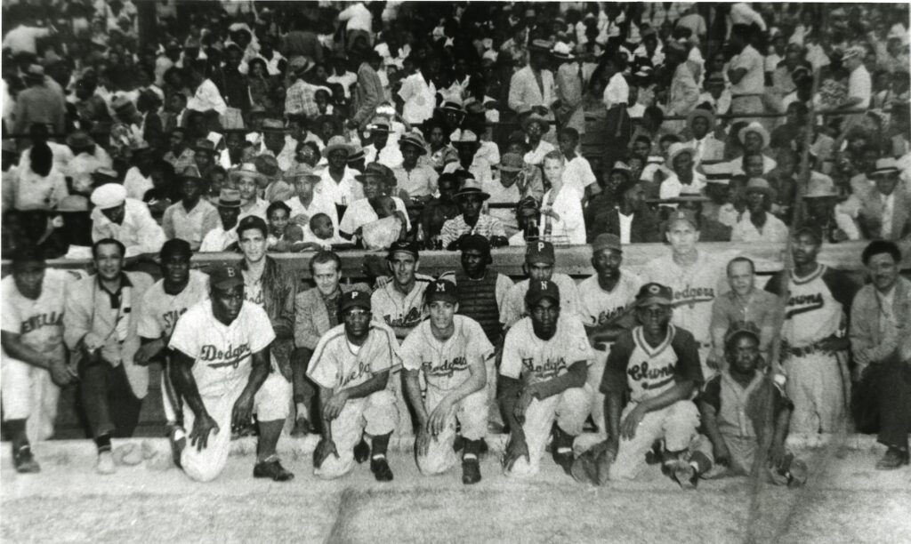 A group of baseball players in vintage uniforms kneel in the foreground, while a large crowd of spectators fills the stadium seats behind them. The atmosphere is lively, with people wearing hats and casual attire typical of a bygone era.