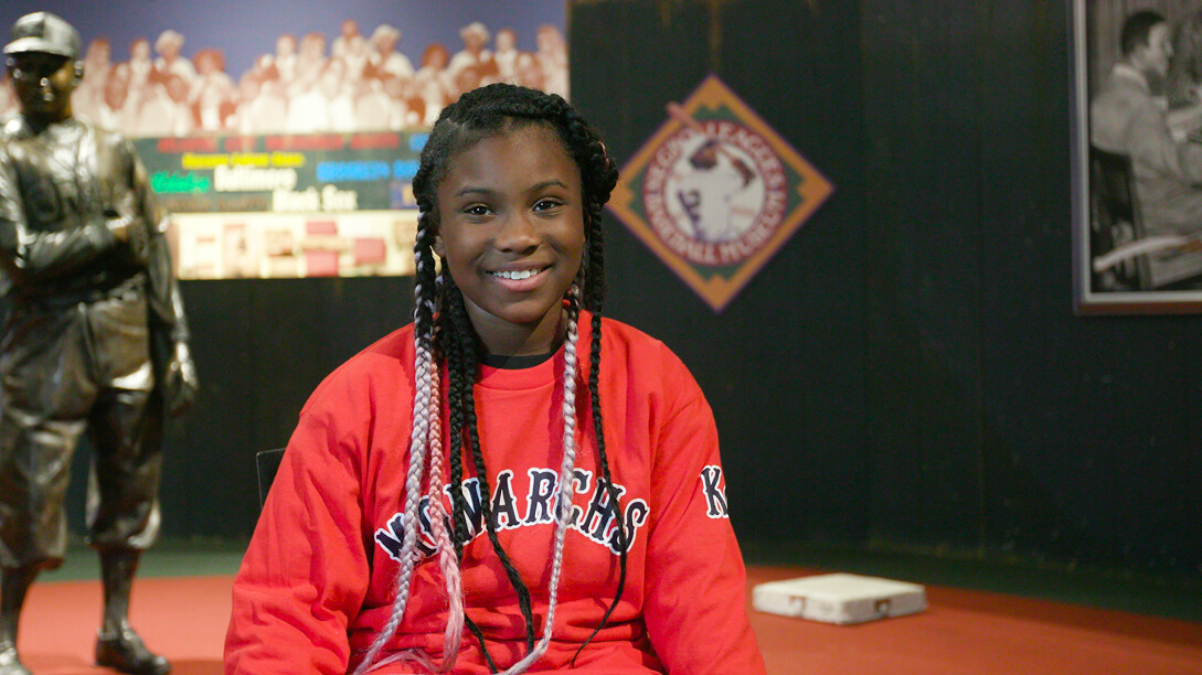 A young person with braided hair, wearing a red shirt that reads Kansas City Monarchs, smiles while seated. Behind them, a statue of a baseball player and colorful banners are visible.