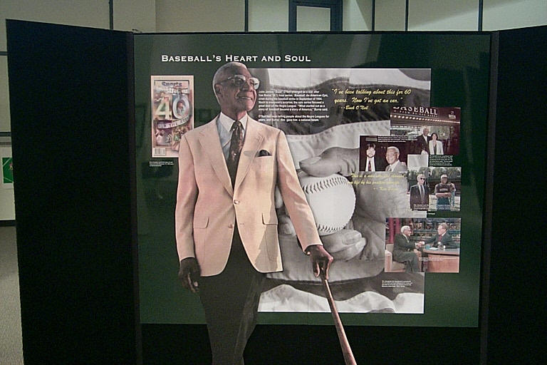 Display board titled Baseballs Heart and Soul featuring an older man in a light-colored suit holding a bat. Background includes various photos and text related to baseball.