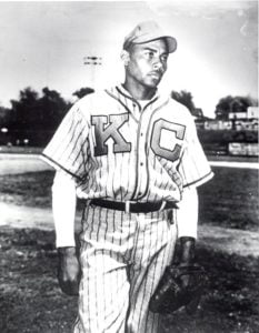 A baseball player wearing a vintage Kansas City Monarchs uniform stands on the field. The uniform is pinstriped with K C on the chest. He holds a glove and wears a cap, with a stadium in the background. The photo is in black and white.