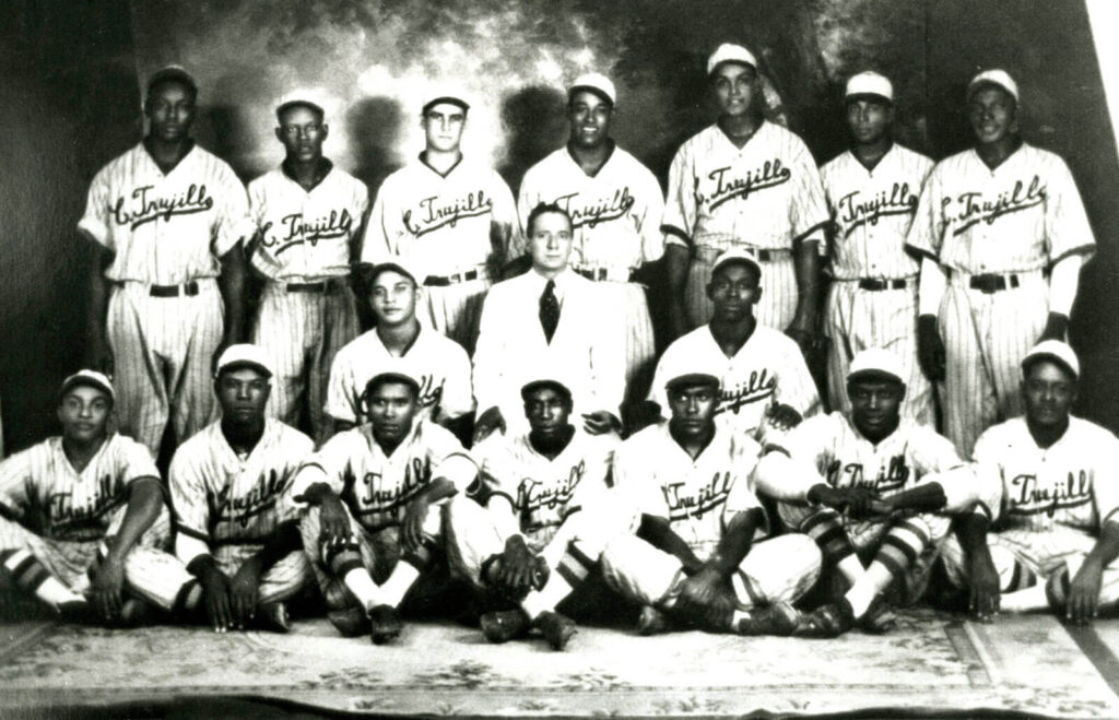 Black and white photo of a baseball team, featuring eighteen men in striped uniforms and caps, and one man in a suit at the center. The team is arranged in three rows, with some standing and others seated.