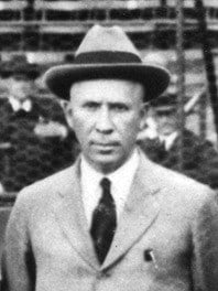A historical black-and-white photo of a man wearing a suit, tie, and fedora hat. The background features a blurred crowd and fence, suggesting an outdoor event or gathering. The man has a serious expression.