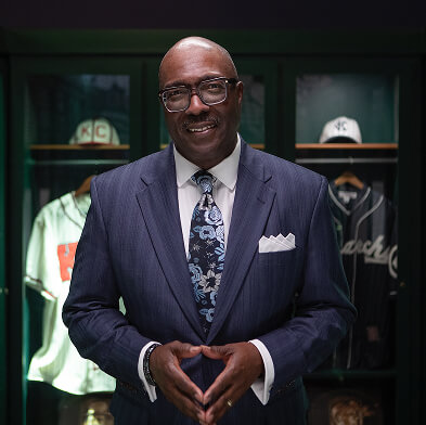 A man in a blue suit and floral tie stands confidently with his hands forming a triangle. Behind him is a display of vintage baseball jerseys and caps in a dark-lit room.