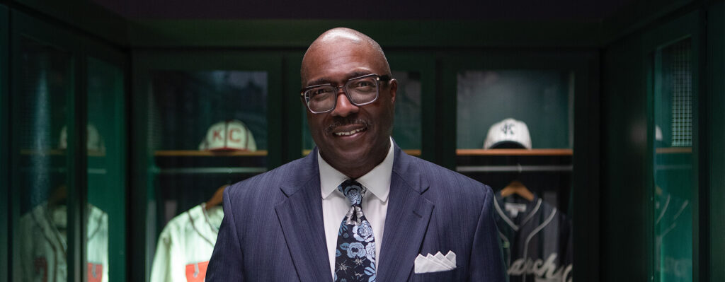 A man in a suit and floral tie is smiling, standing in front of a display case with vintage baseball jerseys and caps. The room is dimly lit with a focus on the man.