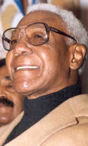 Close-up of a smiling elderly man with white hair, wearing glasses, a brown coat, and a black turtleneck. A blurred image of another person appears in the background.