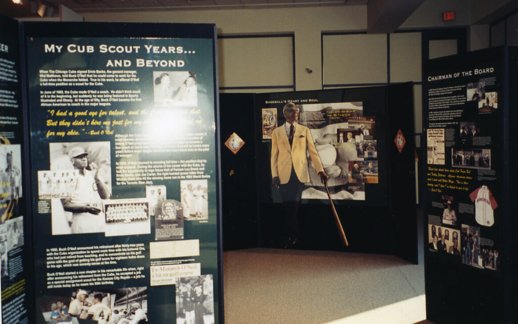 Museum exhibit featuring panels about personal histories and achievements, including photographs and text. Central panel shows a person in a yellow jacket holding a baseball bat.