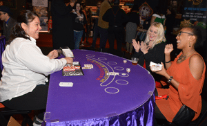 Three people at a casino-themed table, with a dealer on the left and two women on the right, smiling and clapping. There are playing cards and chips on the table, with a background of people and casino decor.
