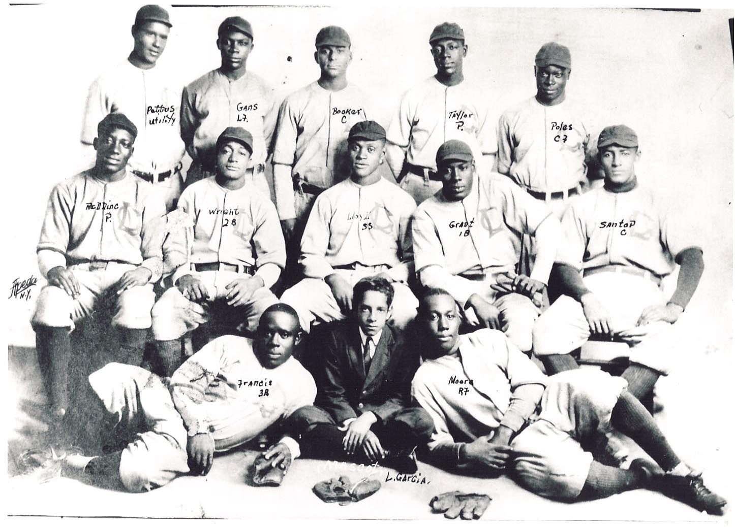 Historic baseball team group photo. Thirteen players posing in uniforms with some names written on the image. Three players sitting, two lying in front with a suited man between them. Baseball gloves placed on the ground. Black and white photo.