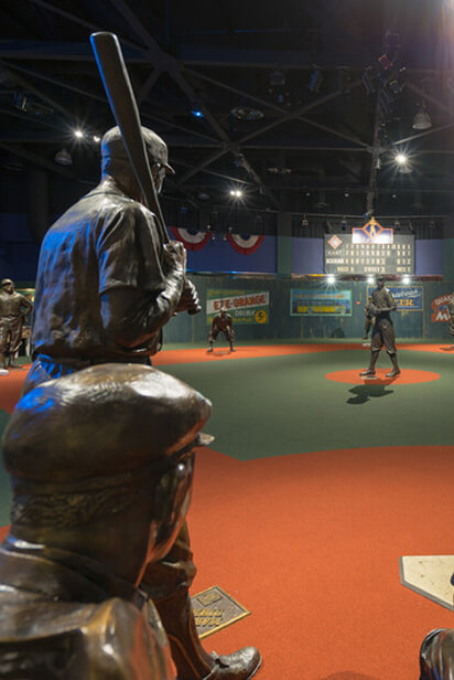 Bronze statues depicting a baseball game scene on a red and green field. A batter stands ready to swing as a pitcher prepares to throw the ball. The background features vintage advertisements and a scoreboard under a dark ceiling with lights.
