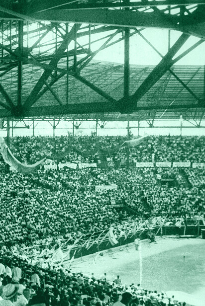 A large stadium filled with spectators. The people are seated in tiered rows under a metal roof with visible beams. The scene has an old, vintage feel, as the image is tinted green. Banners and flags are hanging among the crowd.