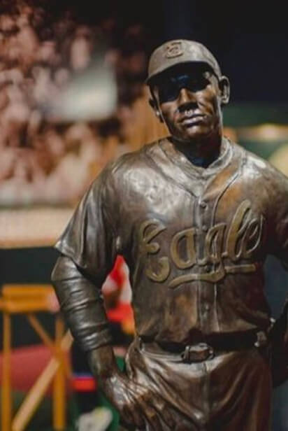Bronze statue of a baseball player wearing a cap and a jersey with Eagle written on it. The player is posed with one hand on their hip. The background is blurred, featuring some indistinct objects and structures.