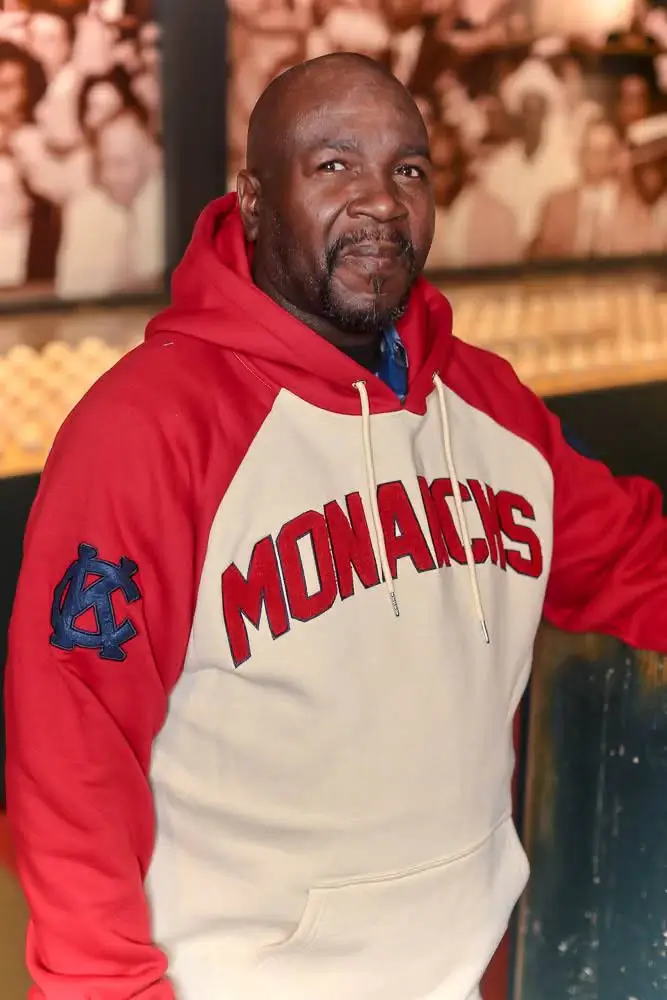 Man sitting wearing the Homestead Grays Heritage Hoodie, featuring a white body with a red Monarch's logo on the front, red sleeves and good, with a blue KC mark on the shoulder