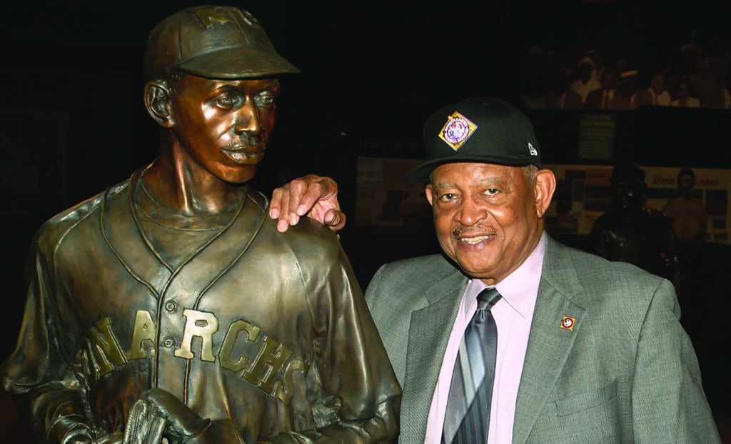 An elderly man in a suit and baseball cap smiles while standing next to a bronze statue of a baseball player wearing a Monarchs jersey. The man rests his hand on the statues shoulder.