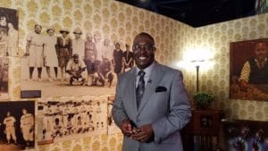 A man in a gray suit stands smiling in a room with vintage photographs on the walls. The background features old sepia-toned images and a vintage lamp, creating a historical atmosphere.