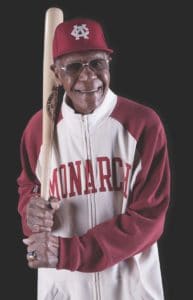 A person wearing a red and white baseball uniform and cap, labeled Monarch, stands smiling while holding a baseball bat on their shoulder. The uniform has a vintage style, and the background is dark.