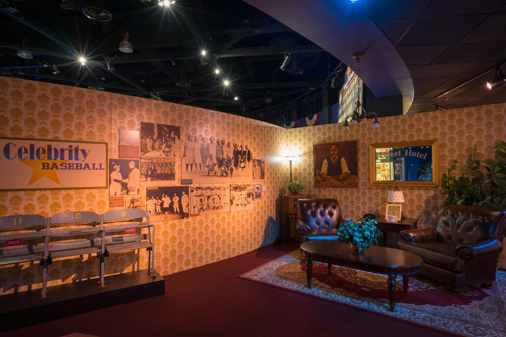 A cozy museum exhibit features vintage baseball memorabilia. It includes a Celebrity Baseball sign, historic photos, a row of stadium seats, leather chairs, a coffee table, plants, and warm lighting creating an inviting atmosphere.