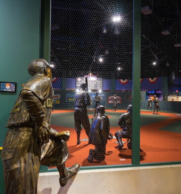 Statues of baseball players in action pose on a red carpeted display surrounded by netting, with dim lighting and baseball-themed decorations in the background.