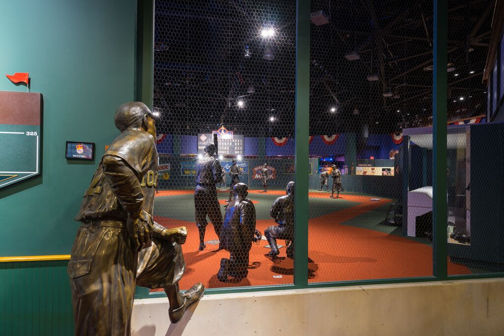 A museum exhibit with baseball player statues staged in action scenes. A statue of a player, wearing a uniform, leans on a railing, observing the scene behind a net. The room is dimly lit with sports memorabilia on display.