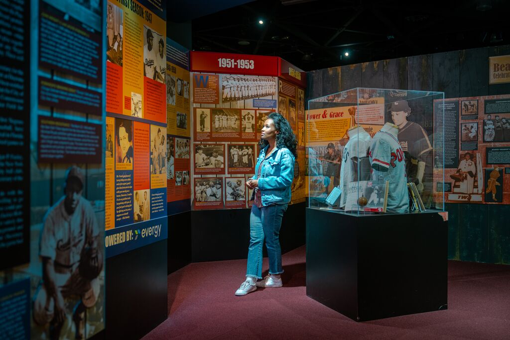 A person in a blue jacket and jeans stands in a museum exhibit filled with historical baseball memorabilia, including jerseys and photographs from 1951-1953. They are observing the displays closely, surrounded by colorful information panels.
