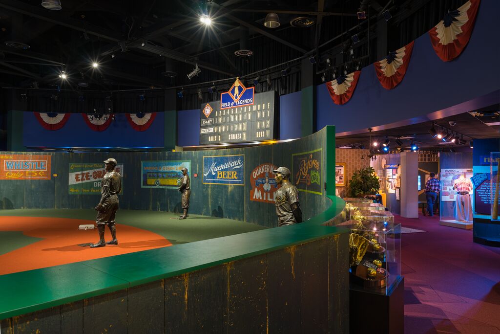 A museum exhibit features a small baseball field with life-sized statues of baseball players. Colorful vintage advertisements and a scoreboard hang on the walls. Display cases and decorative lights are seen in the background.