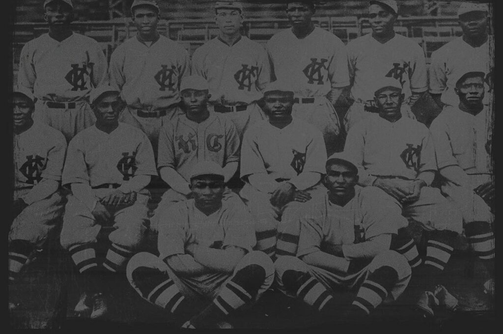A vintage black-and-white photo of a baseball team, with 15 players in uniforms and caps, sitting and standing in rows on a field. The uniforms feature a distinct logo on the chest. The background shows empty stadium seating.