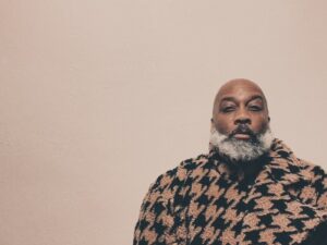 A person with a graying beard is wearing a black and brown houndstooth coat. They are positioned slightly to the right against a plain, light-colored background. The expression is calm and relaxed.