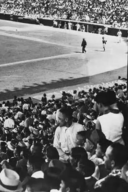 Black and white photo of baseball field, centered on home plate with a baseball player and umpire in view. The crowd is in the foreground and background.