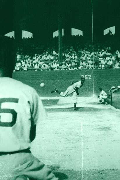 view of player throwing ball from home plate during a game, with fans in the stadium, the back of another player is seen in the foreground