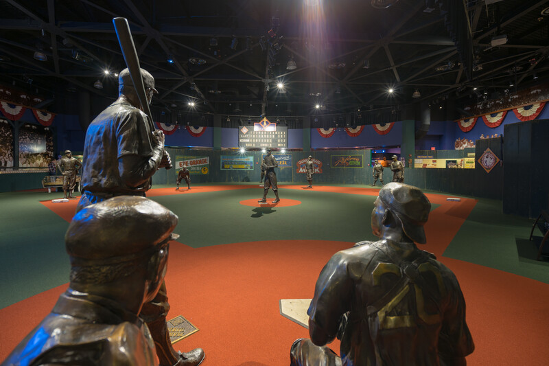 Bronze statues of baseball players are positioned on a replica field inside a museum. The room is decorated with pennants and vintage advertisements. Overhead lighting highlights the scene, creating a dramatic atmosphere.