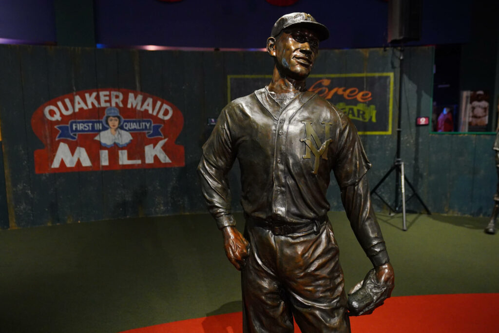 Bronze statue of a baseball player in uniform holding a glove, with vintage Quaker Maid Milk sign in the background.
