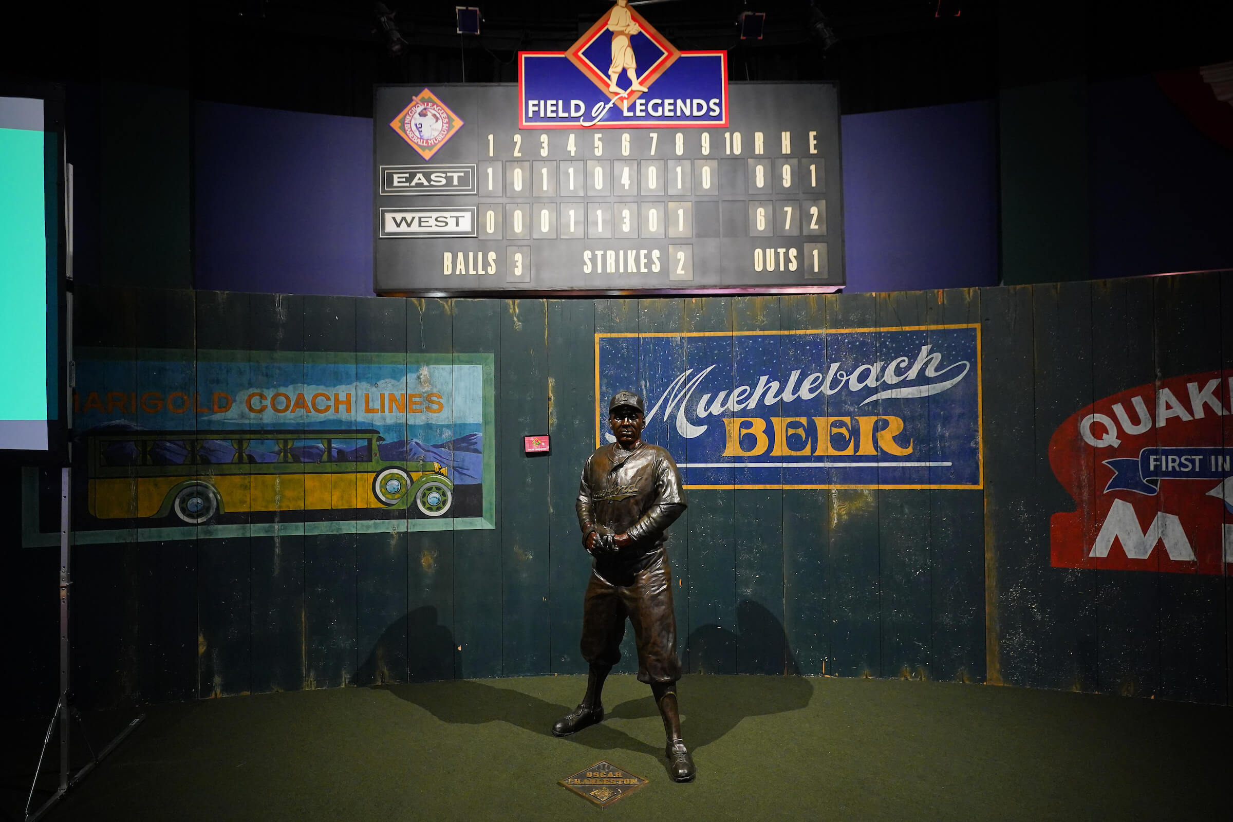 A bronze baseball player statue stands in front of a vintage-style scoreboard, with signs for Muehlebach Beer and Aristo Coach Lines on a green wall backdrop.
