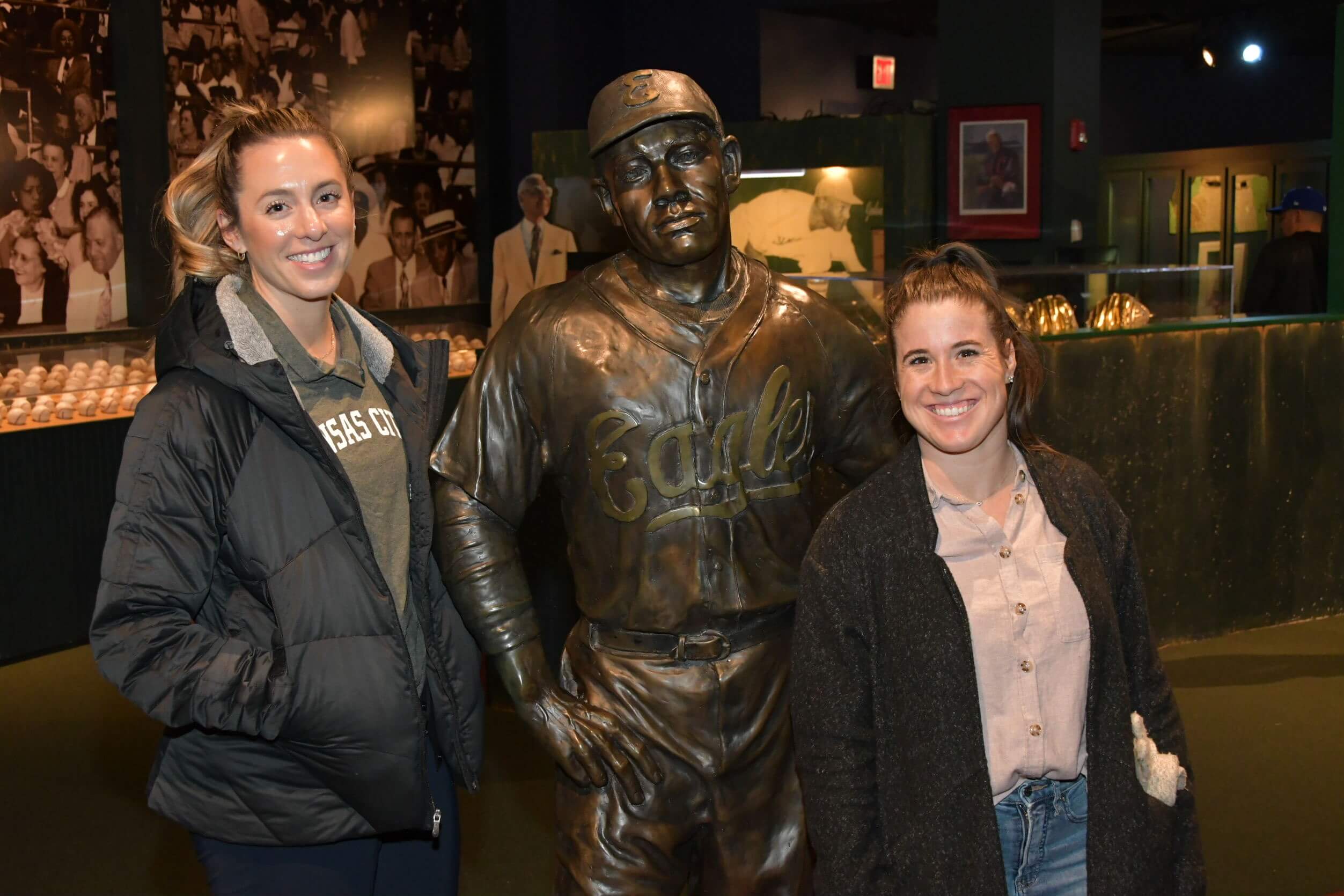Two women stand beside a bronze baseball player statue in a museum. The background features framed photos and a display of baseballs.