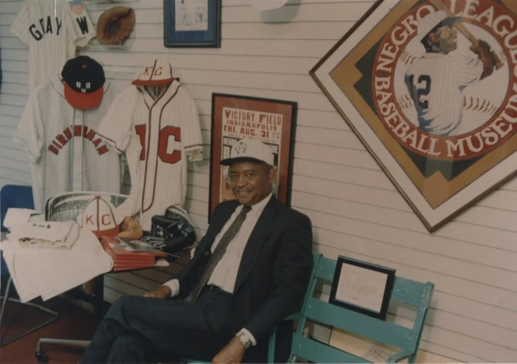 An individual sits at the Negro Leagues Baseball Museum, surrounded by vintage baseball memorabilia, jerseys, and a framed poster on the wall.
