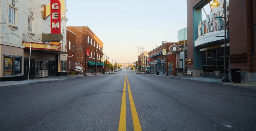 A deserted city street lined with brick buildings, restaurants, and shops. Yellow lines run down the center. A marquee sign reads "GEM" on the left. Bright, calm morning light.