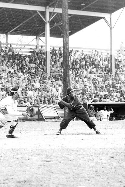 A baseball player swings at a pitch during a game, with a catcher and an umpire in position. A crowd fills the stands in the background.