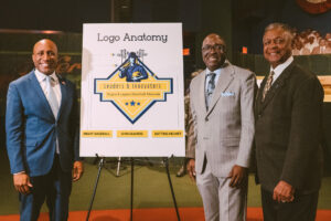 Three men in suits stand beside a poster titled "Logo Anatomy" at an event for the Negro Leagues Baseball Museum.